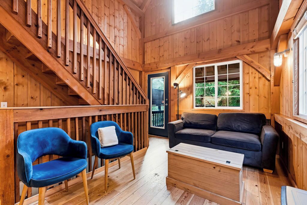 A wooden interior with a staircase, two blue chairs, a black sofa, and a wooden coffee table.