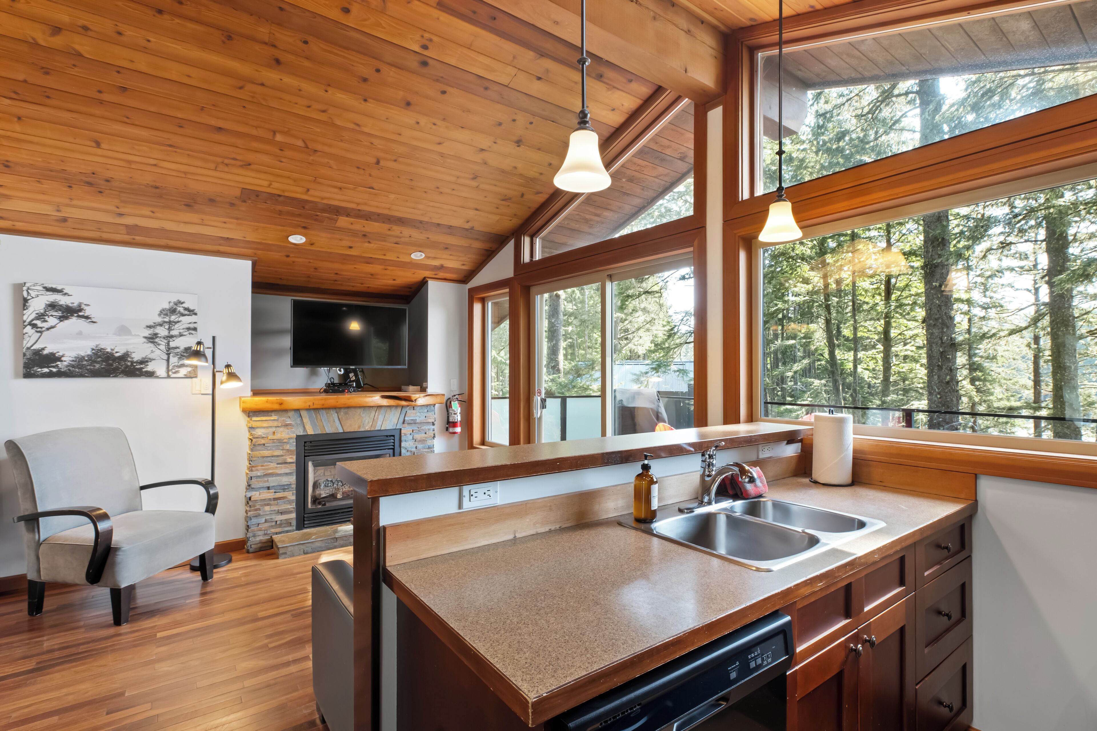 A modern kitchen with a stone fireplace, wooden ceiling, and large windows overlooking a forest.