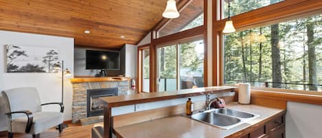 A modern kitchen with a stone fireplace, wooden ceiling, and large windows overlooking a forest.
