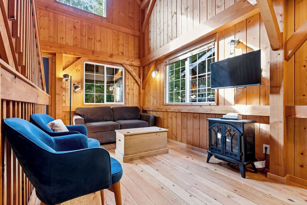 A wooden cabin interior with a sofa, two chairs, a television, and a wood-burning stove.