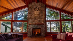 Una habitación acogedora con chimenea de piedra, techo de madera y ventanas amplias con vistas al bosque.
