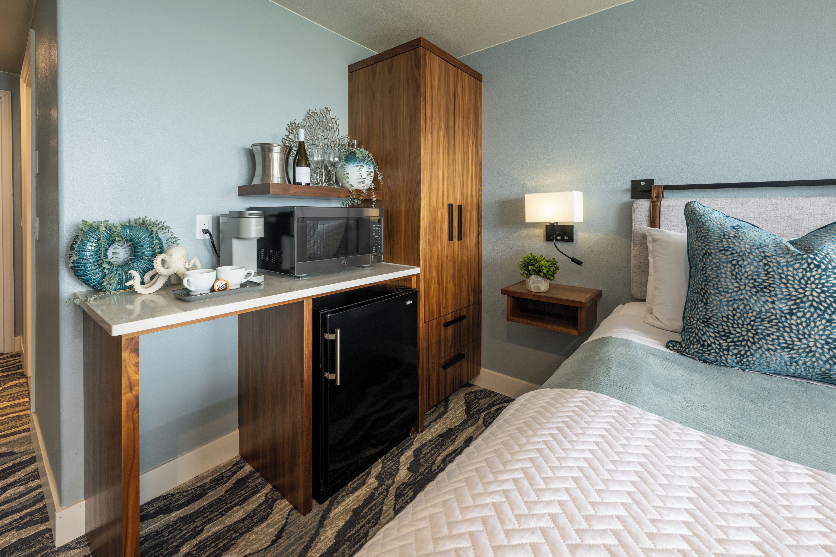 A compact hotel room with a kitchenette, a bed with patterned pillows, and a small bedside table with a lamp.