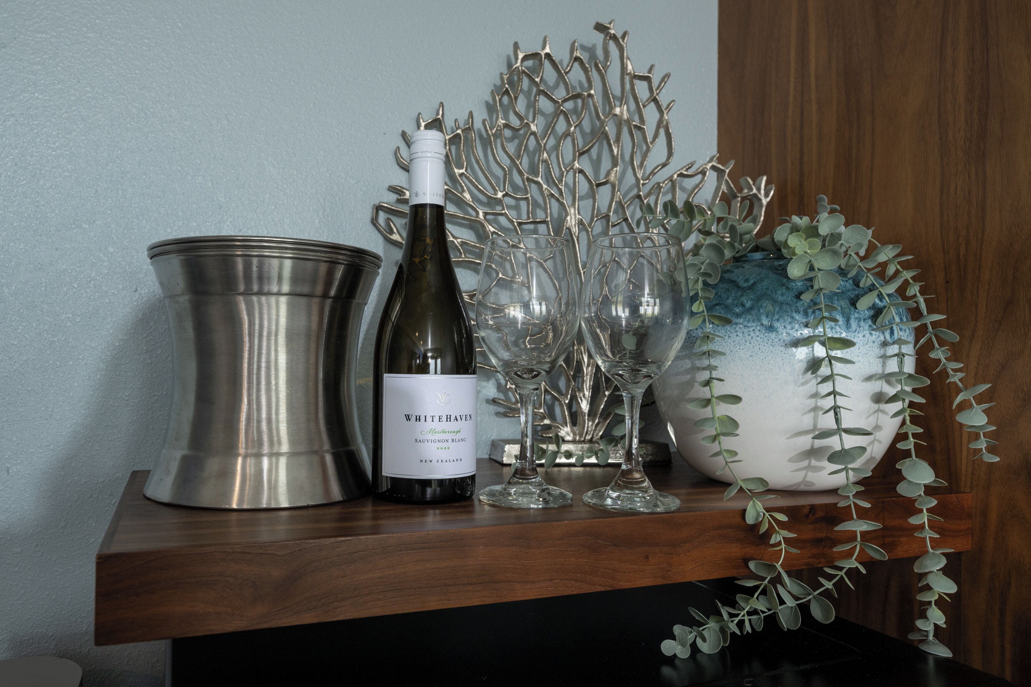 A bottle of Whitehaven Sauvignon Blanc, two wine glasses, a metal bucket, and a ceramic vase with greenery on a wooden shelf.