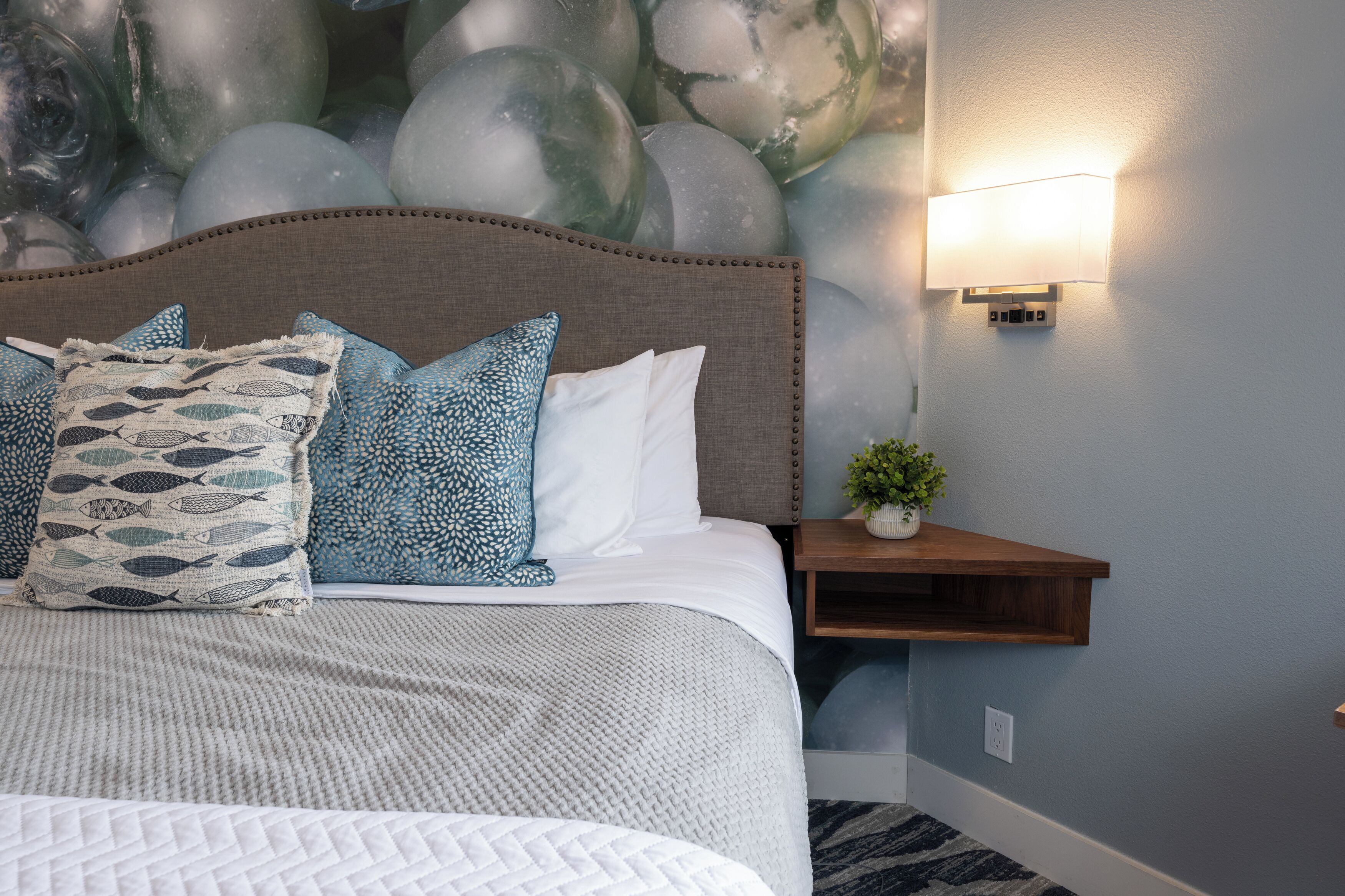 A neatly made bed with patterned pillows, a headboard, a nightstand with a plant, and a wall-mounted light fixture.