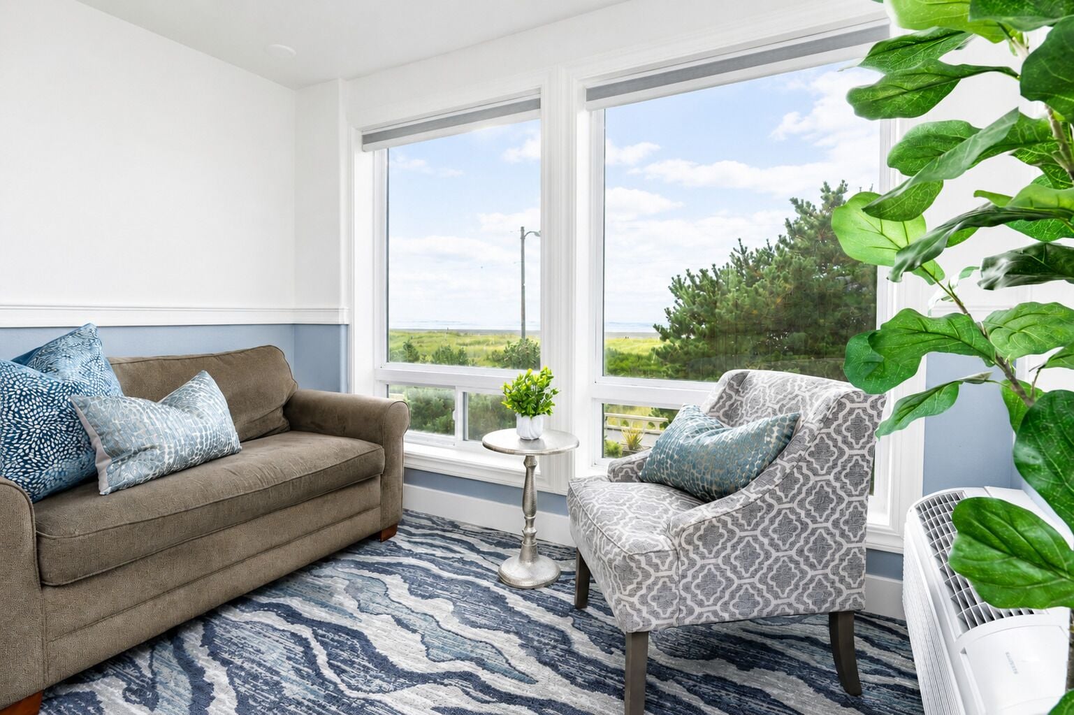 A room with a sofa, armchair, and a view of the outdoors through large windows.
