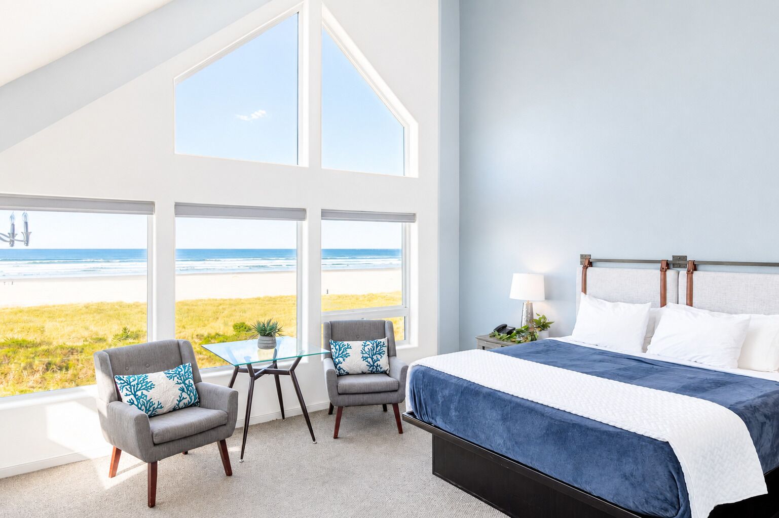 A bedroom with a large window overlooking a beach, a bed with a blue bedspread, two gray armchairs, and a small table.