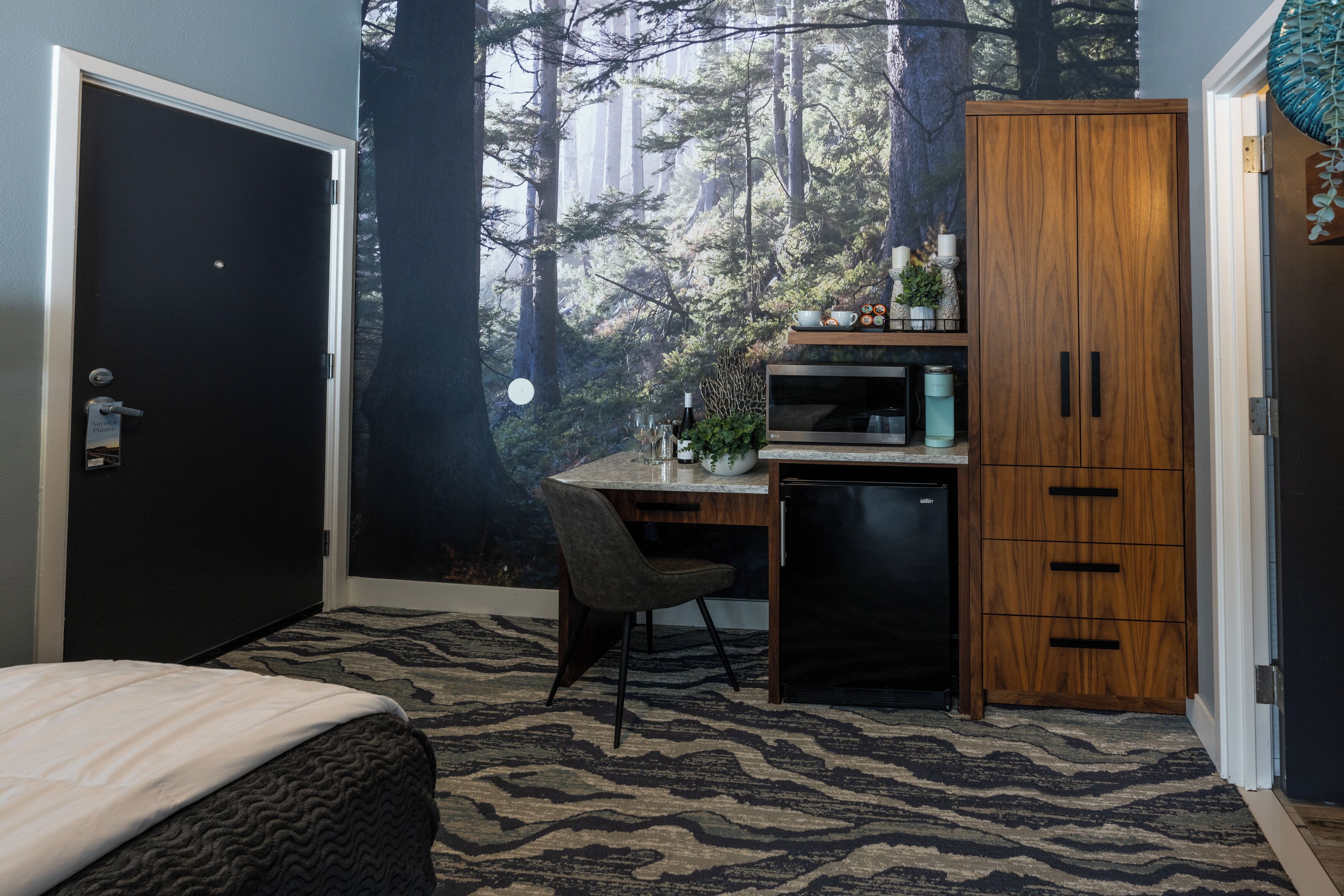 A bedroom with a bed, a desk, a chair, a wardrobe, and a view of a forest through a window.