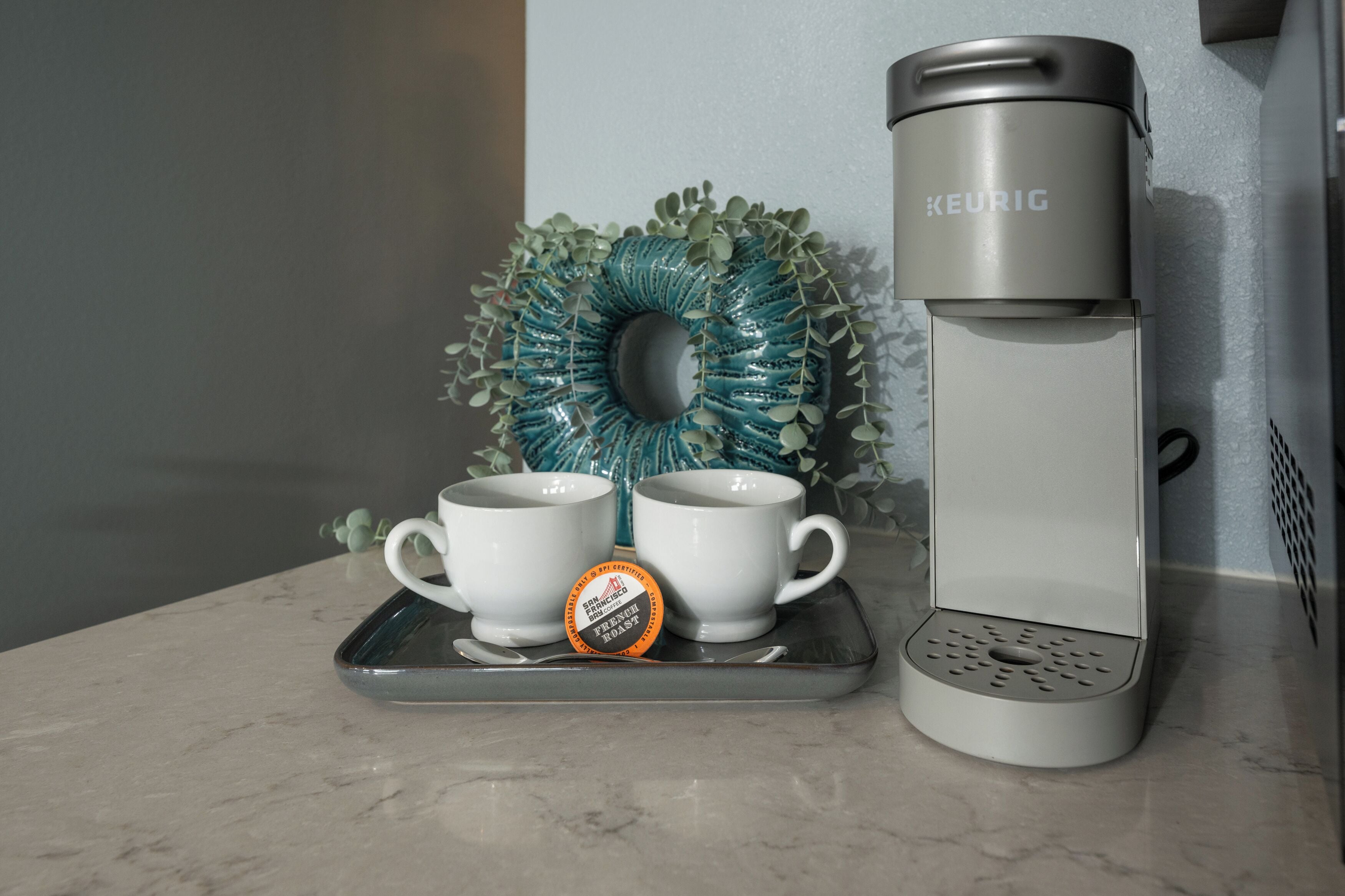 A Keurig coffee maker, two white cups, and a tray with a coffee pod on a marble countertop.