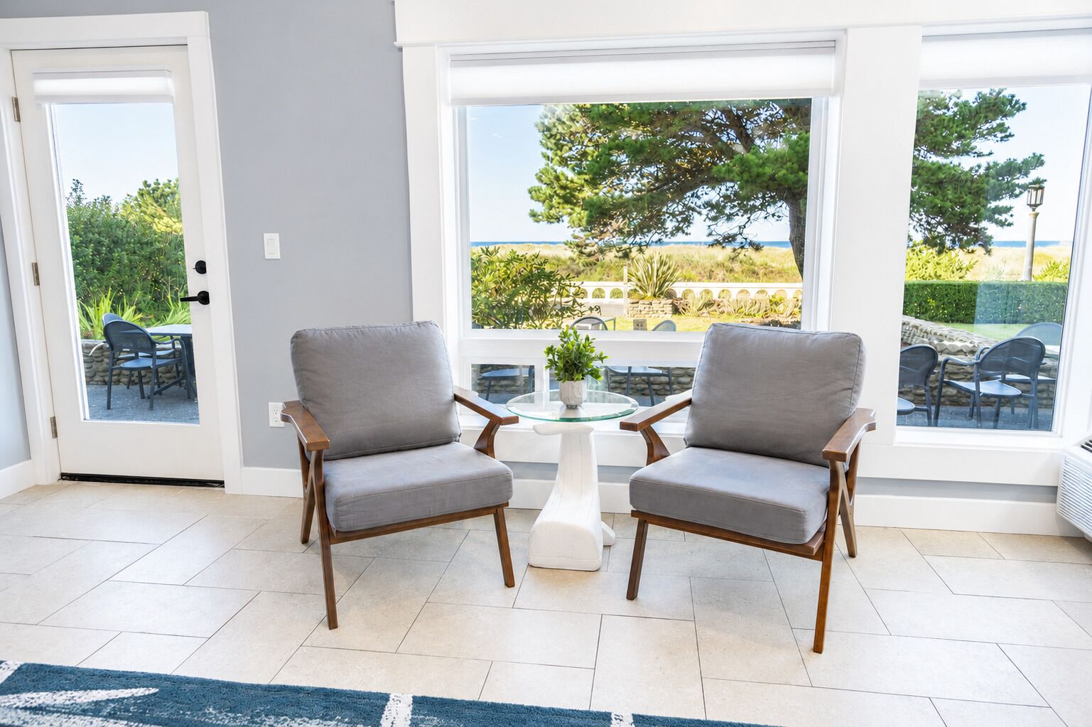 A room with two grey upholstered chairs, a small white table with a potted plant, and large windows offering a view of greenery and a distant landscape.