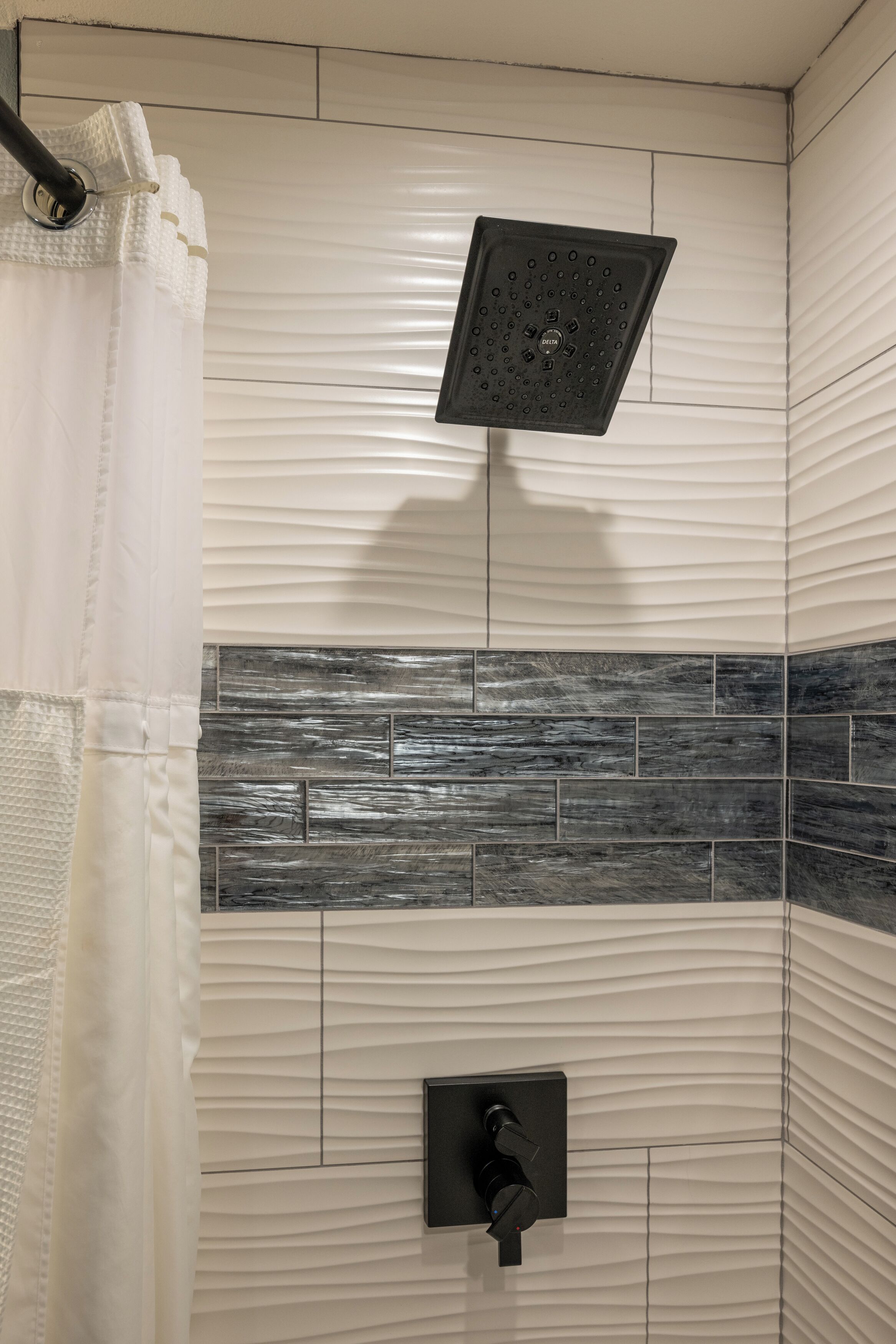 Une douche dotée d’un pommeau de douche noir de type « pluie », d’un porte-serviettes et d’un mur recouvert de carreaux à motifs.