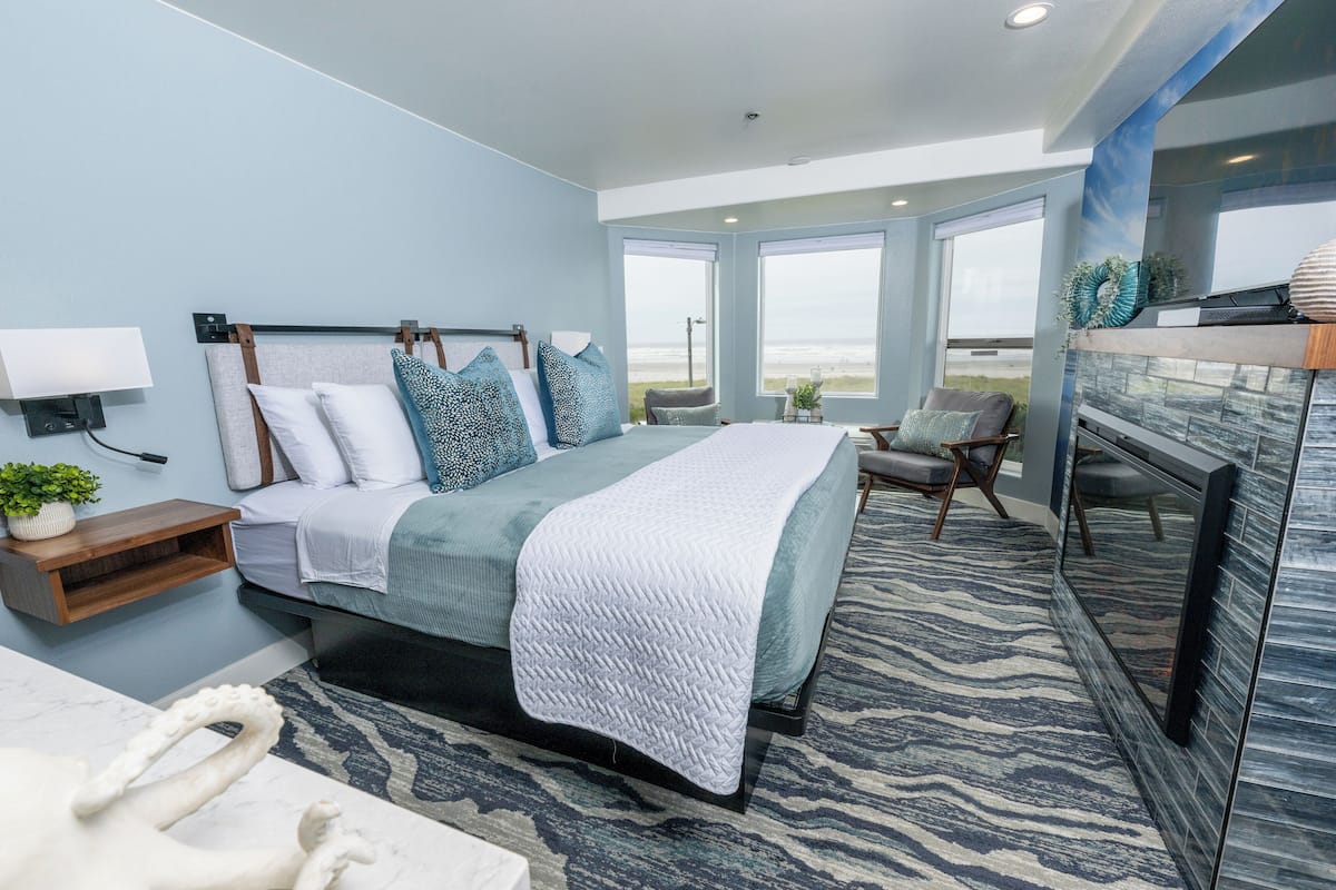 Un dormitorio con una cama grande, una mesita de noche, una silla y vistas a la playa a través de amplias ventanas.
