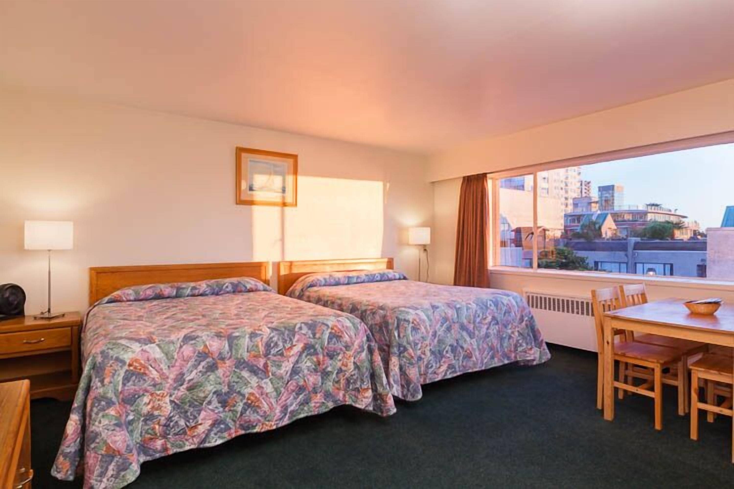 A hotel room with two beds, a dining table, and a view of buildings through large windows.