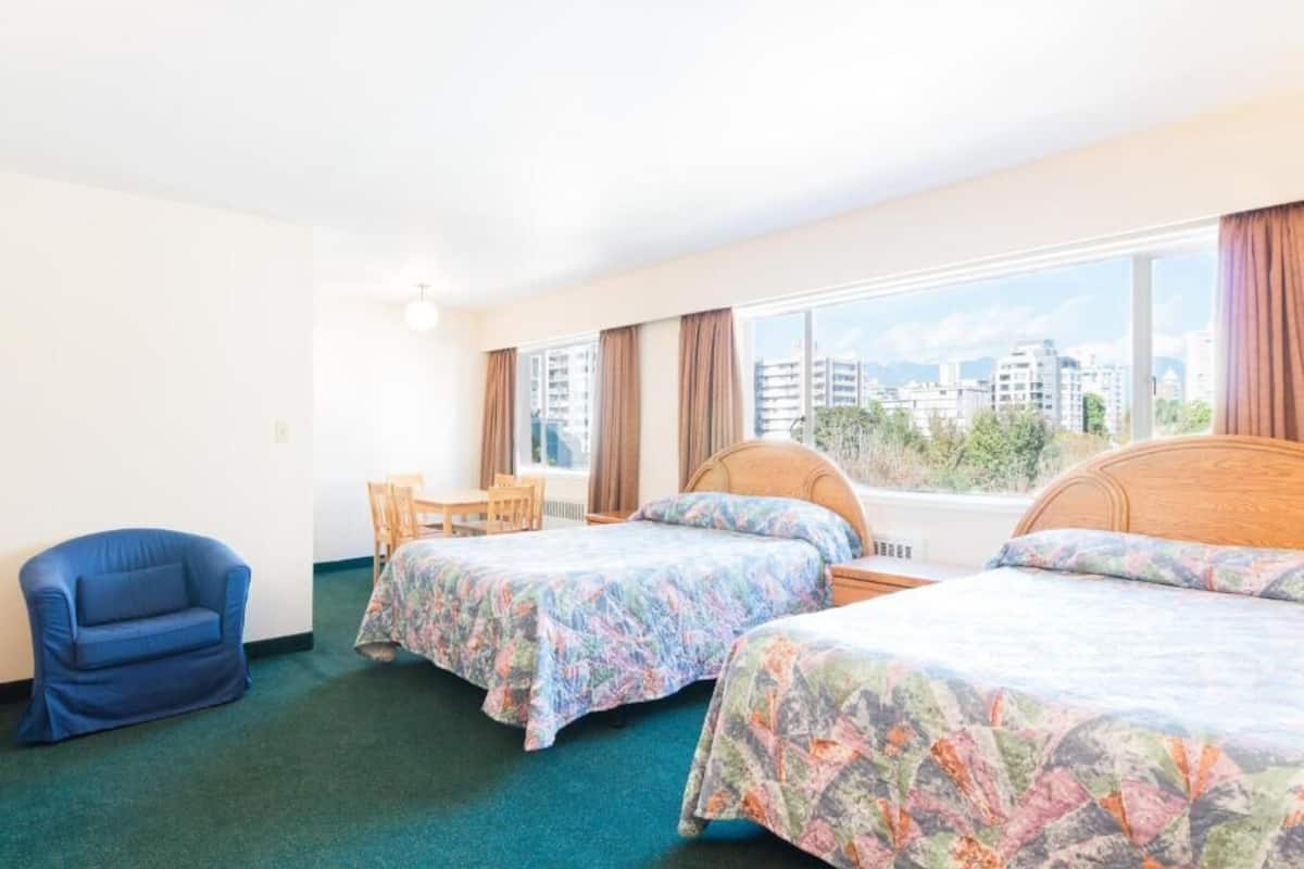 A hotel room with two beds, a blue armchair, a small table, and a window with a city view.