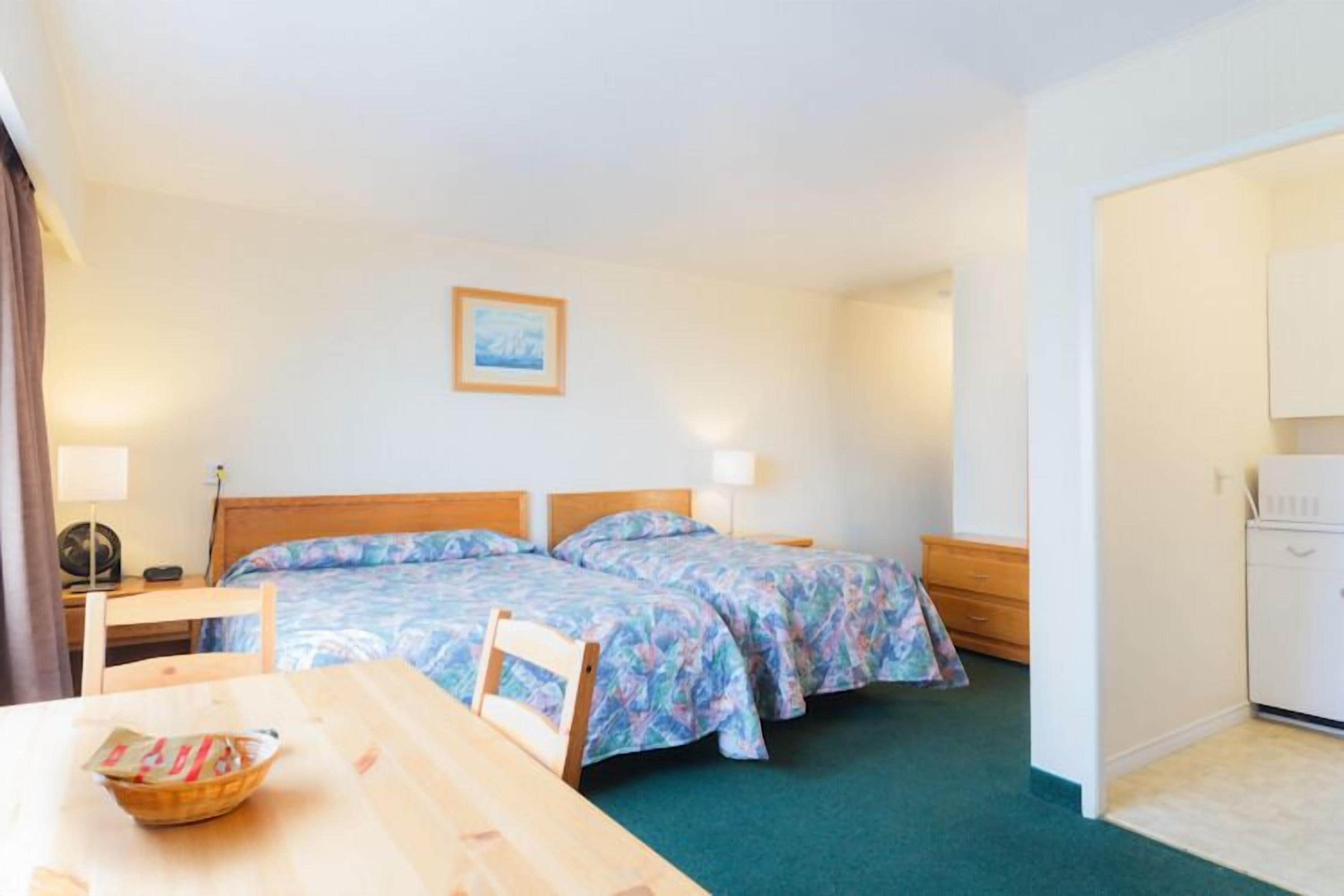 A hotel room with two beds, a wooden desk, a chair, a table with a bowl, a lamp, a picture on the wall, and a white cabinet.