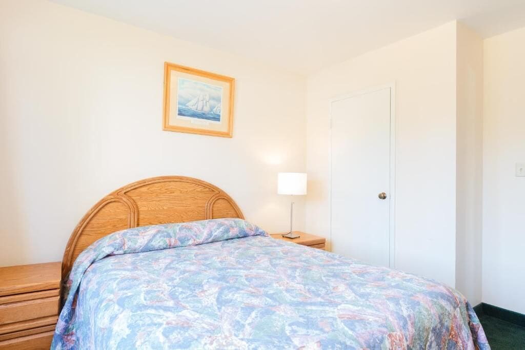 A bedroom with a wooden headboard, a bed with a colorful bedspread, a bedside table with a lamp, and a door.