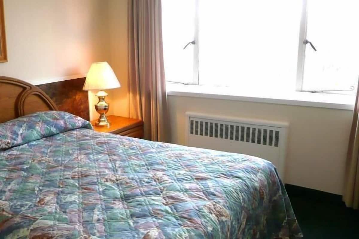 A bed with a patterned bedspread, a bedside lamp, a framed picture on the wall, and a window with curtains.