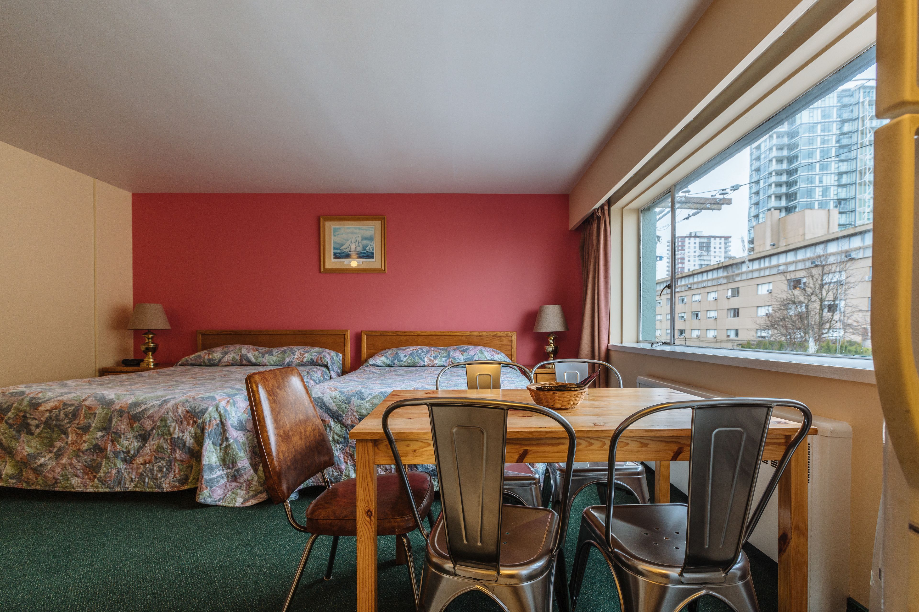 Una habitación con dos camas, una mesa de comedor con sillas, un ventanal con vistas a edificios y un cuadro en la pared.