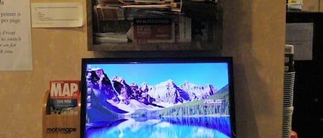 Una computadora portátil muestra un paisaje montañoso, un tablón de anuncios con varios folletos de viajes y un letrero que indica la disponibilidad de una impresora.