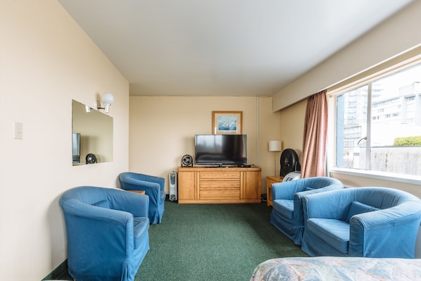 Habitación de hotel con televisión sobre un mueble de madera, dos sillones azules, una mesita con lámpara y una ventana con cortinas.