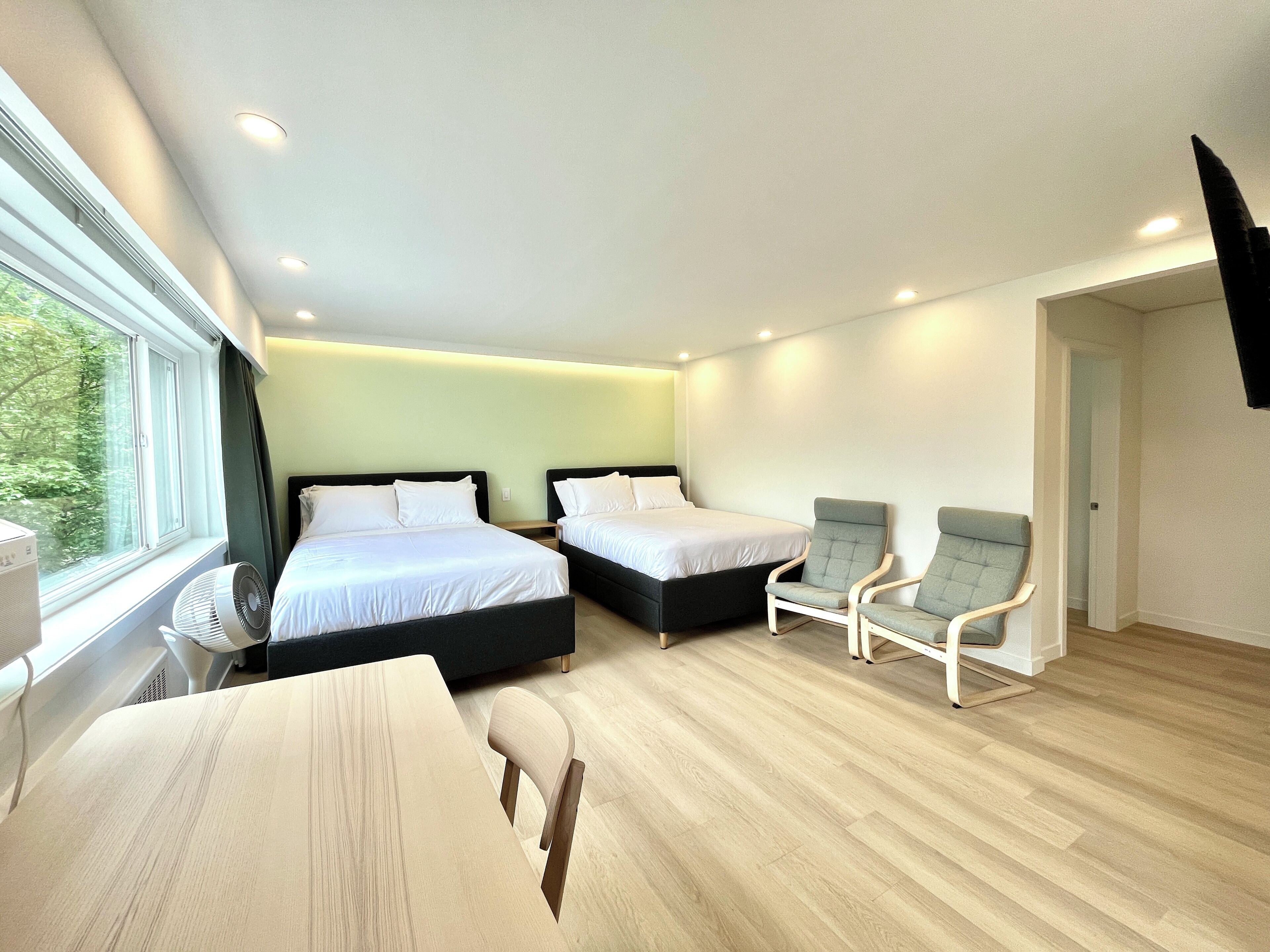 Una habitación de hotel moderna con dos camas, un escritorio, dos sillas, un televisor de pantalla plana y un ventanal con vistas a árboles.