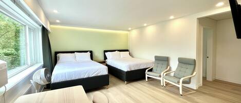 A modern hotel room with two beds, a desk, two chairs, a flat-screen TV, and a window with a view of trees.