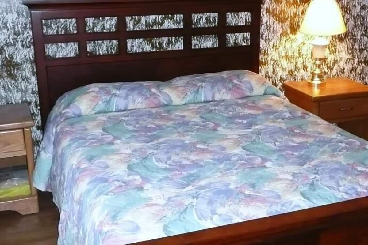 A bed with a floral bedspread, a wooden headboard with glass panels, a nightstand with a lamp, and a patterned wallpaper.