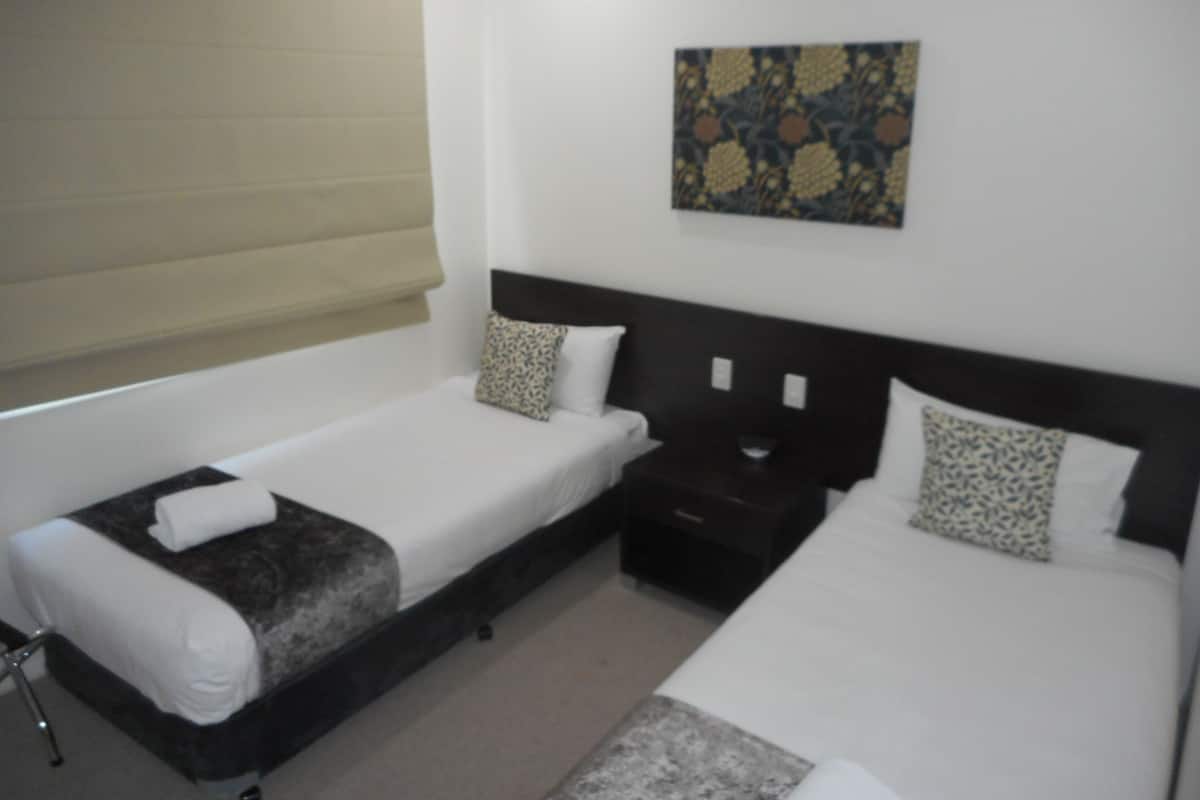 Habitación de hotel con dos camas individuales, una mesita de noche, una pequeña mesa, una ventana con persianas y una obra de arte colgada en la pared.