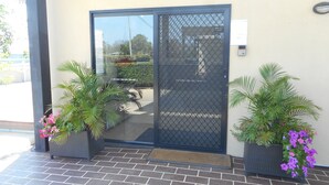 A building entrance with a black metal gate, potted plants with green foliage and pink flowers, and a paved entrance area.