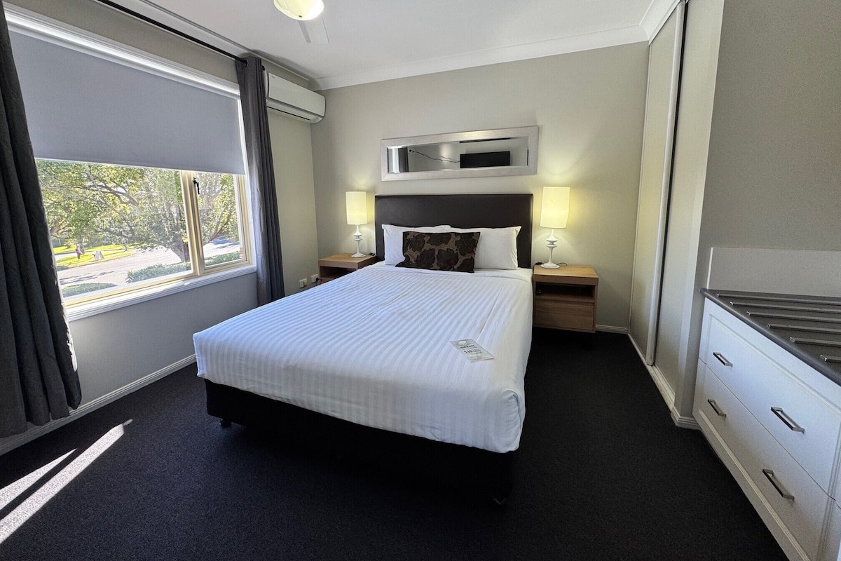 A hotel room with a large bed, two bedside tables with lamps, a ceiling fan, a window with curtains, and a view of the outdoors.