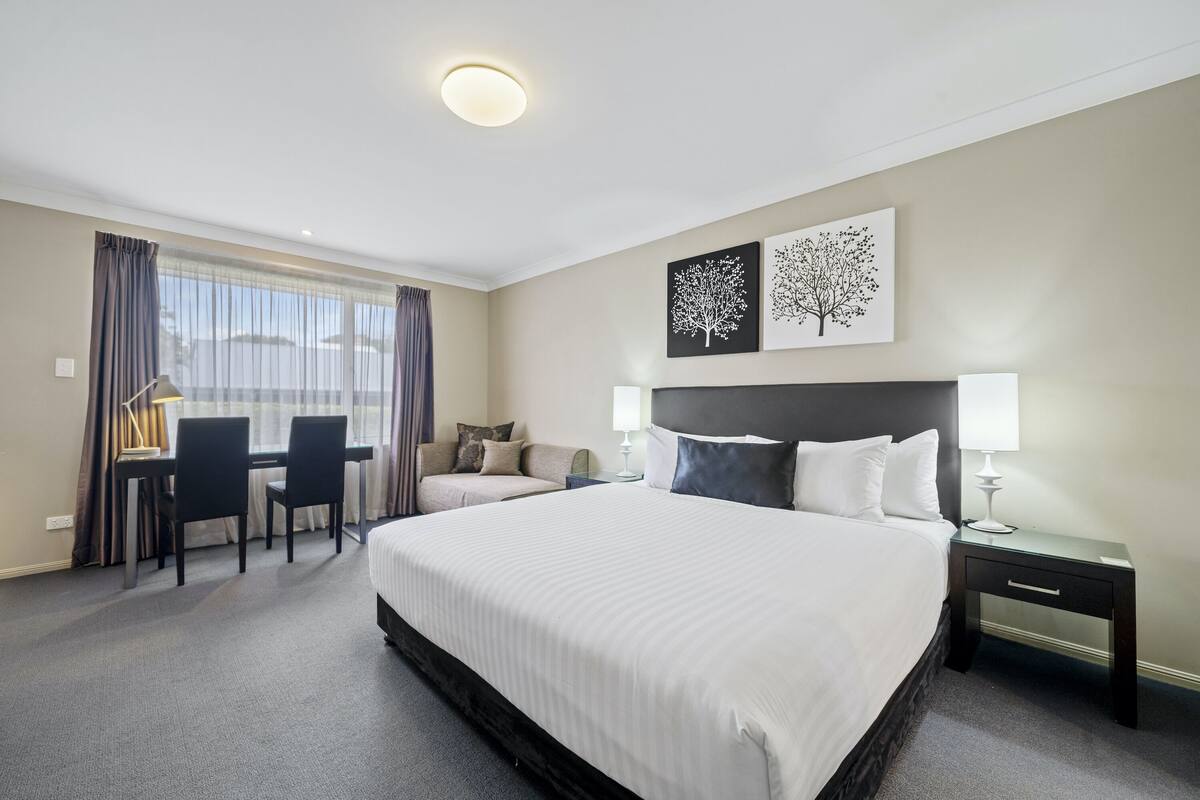 A hotel room with a large bed, a desk with chairs, a nightstand, and two wall art pieces.