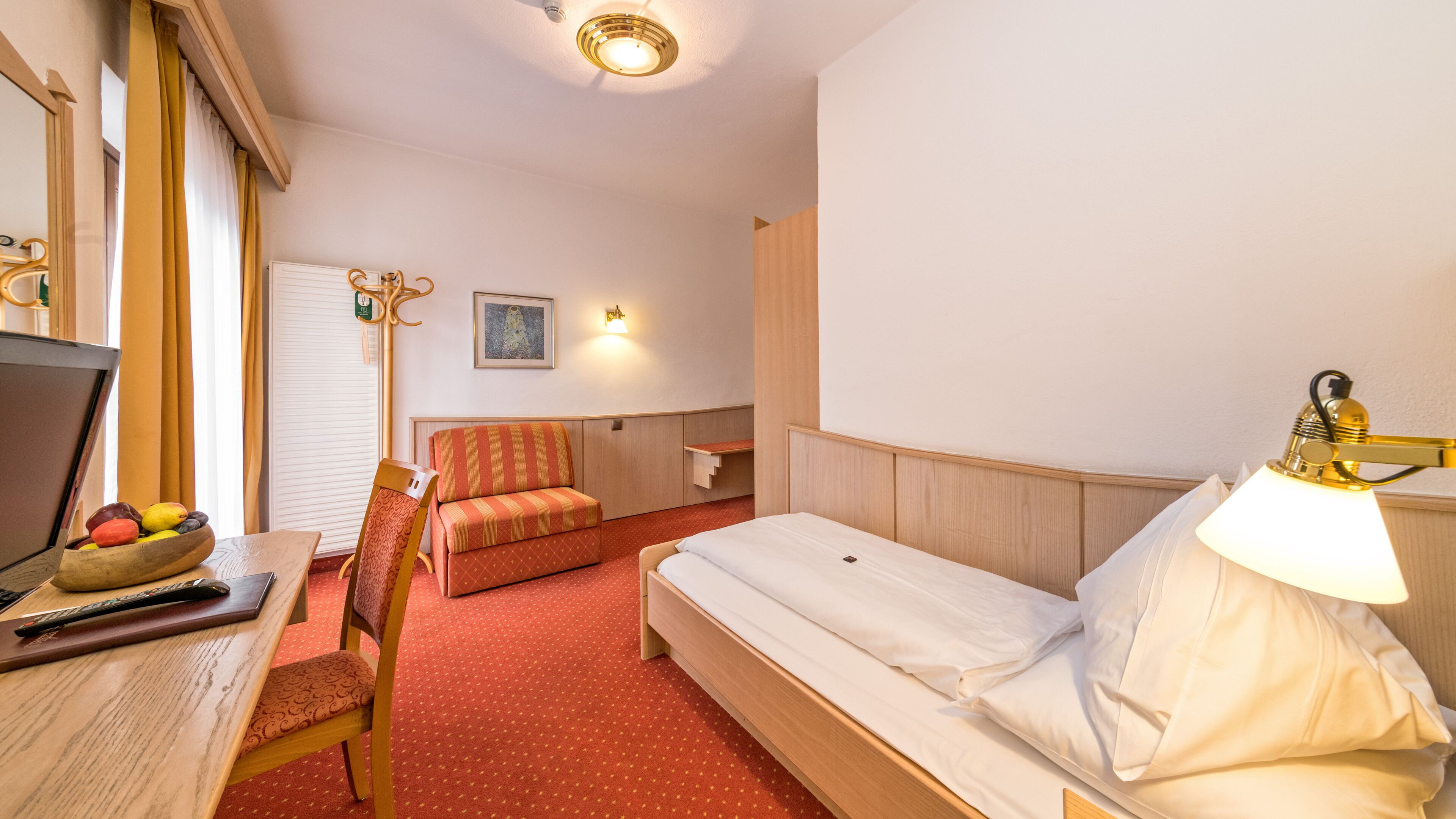 A hotel room with a bed, a desk with a bowl of fruit, a chair, a lamp, and a painting on the wall.