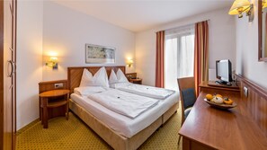 Quarto de hotel com cama de casal, mesa de cabeceira, cadeira, escrivaninha com computador e janela com cortinas.