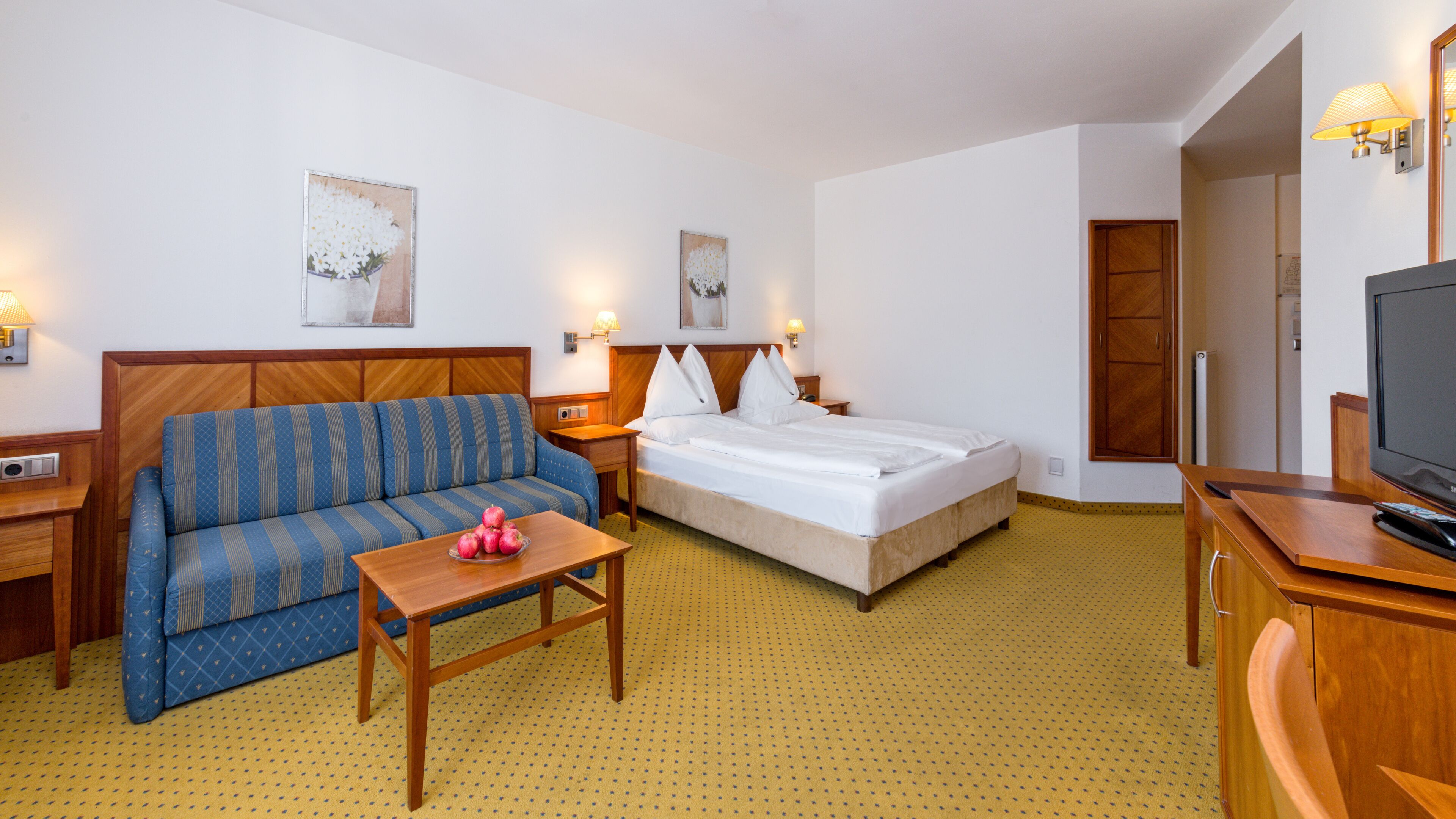 Ein Hotelzimmer mit Bett, Sofa, einem kleinen Tisch, einem Fernseher und Kunstwerken an den Wänden.