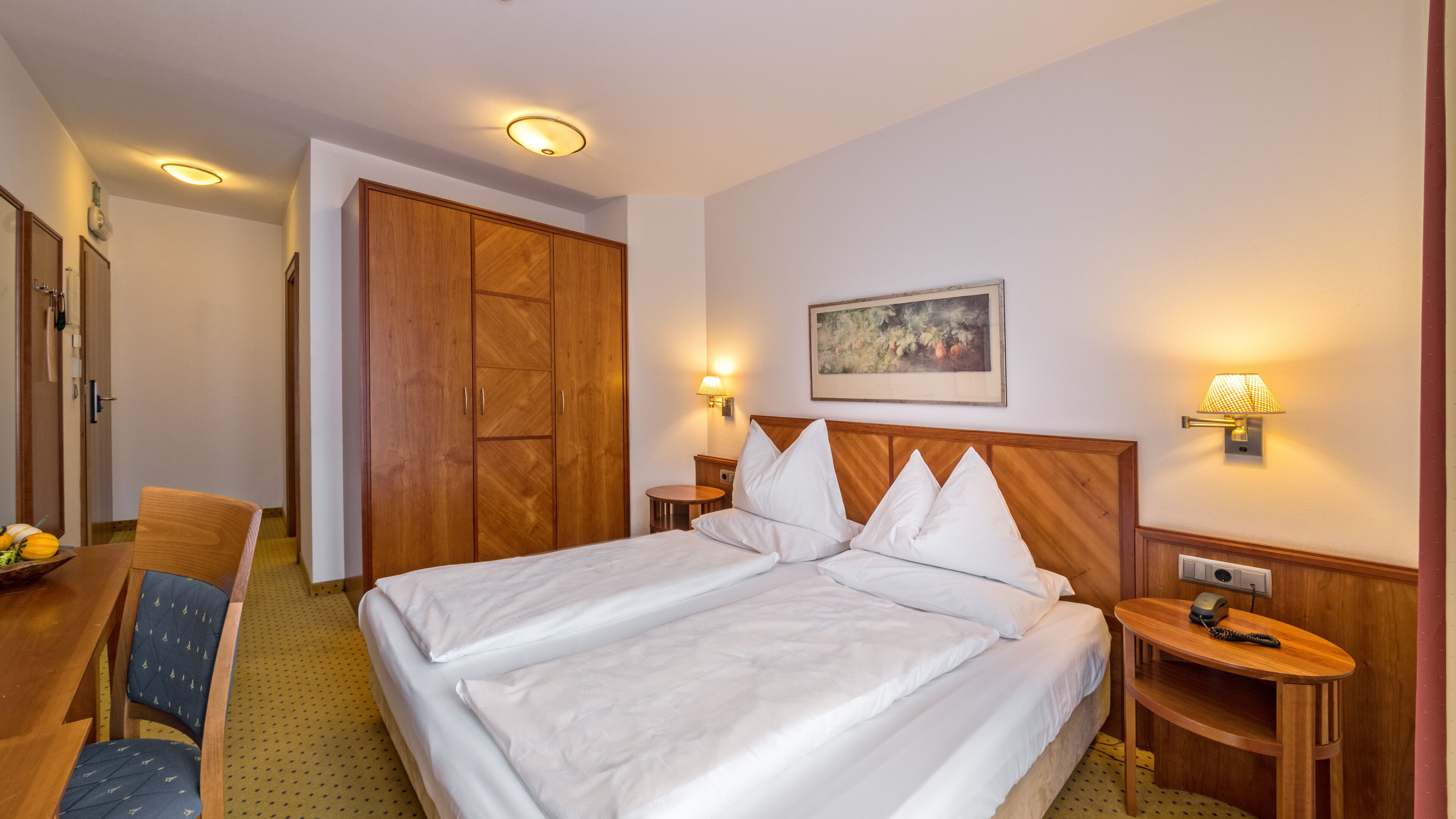 A hotel room with a double bed, wooden wardrobe, dining table, chairs, and a small round table with a lamp.