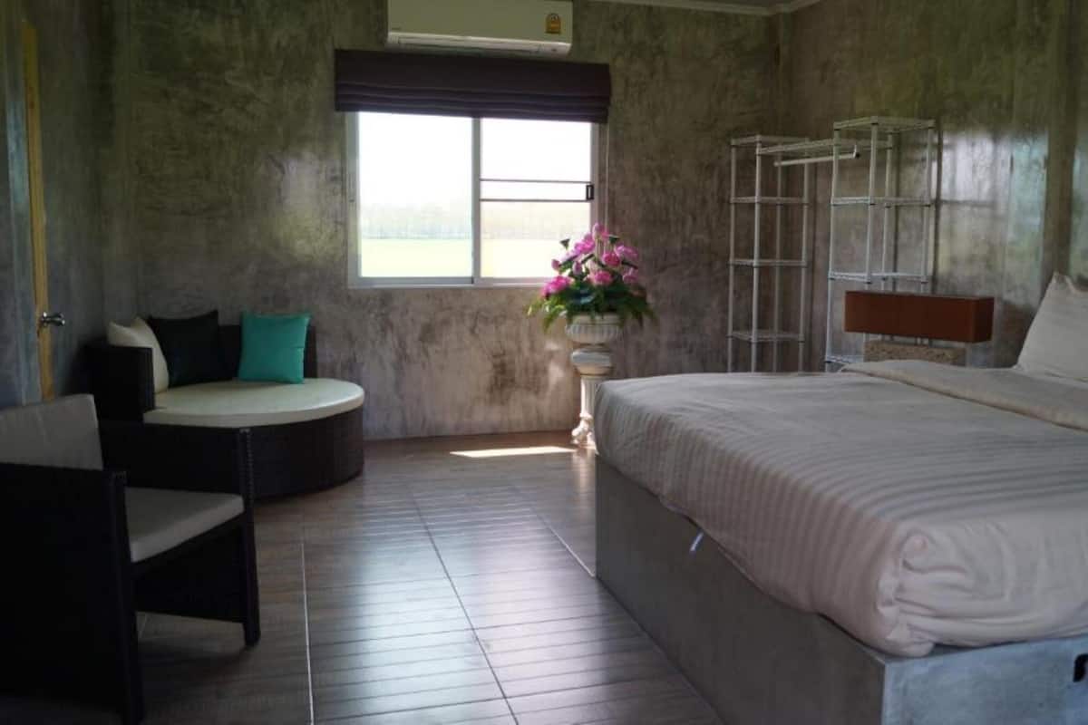 Un dormitorio con una cama grande, una zona de estar circular con un cojín, una ventana con vistas y una estantería metálica.