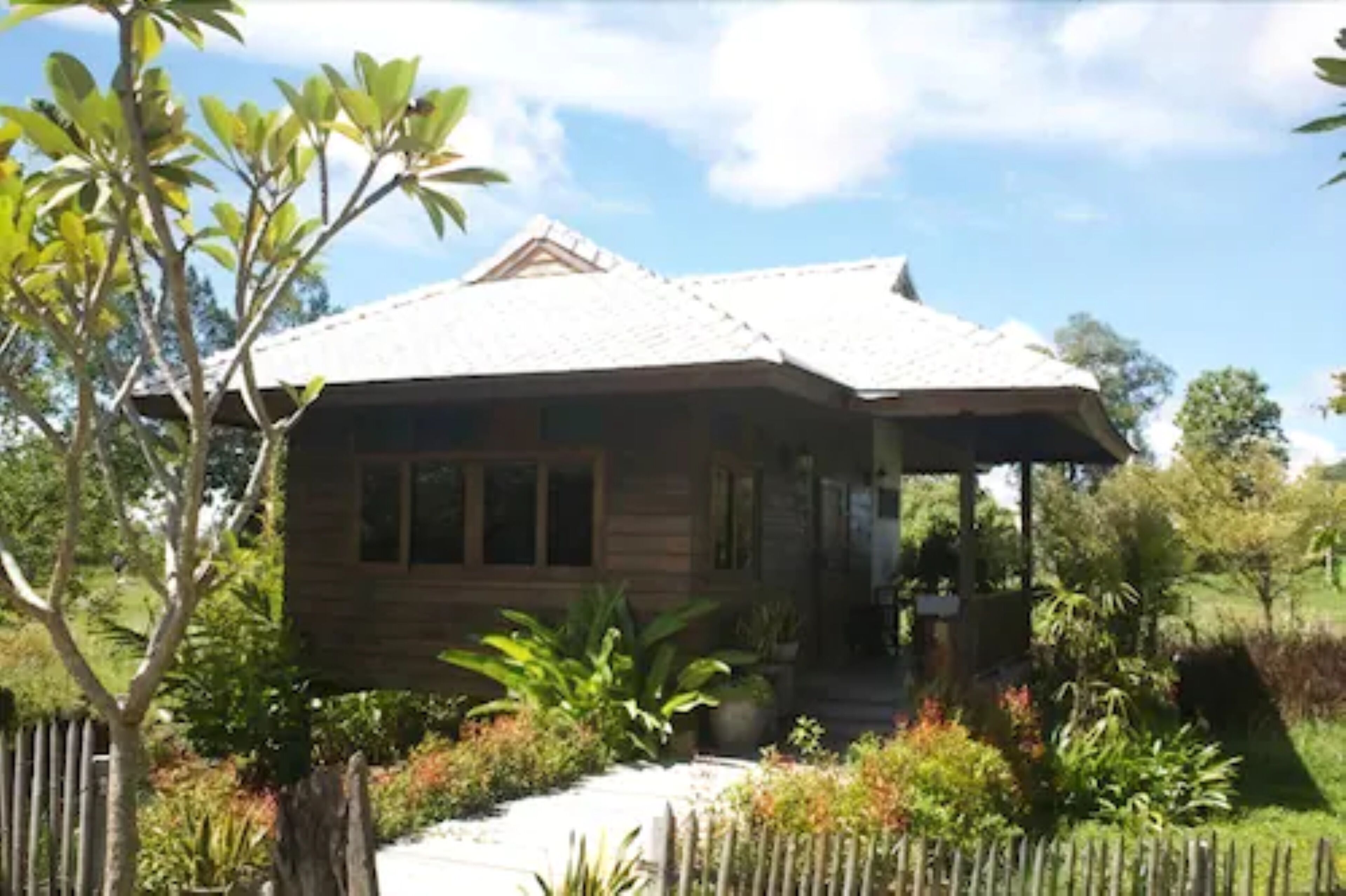 A wooden house with a tiled roof, surrounded by lush greenery and a wooden fence.