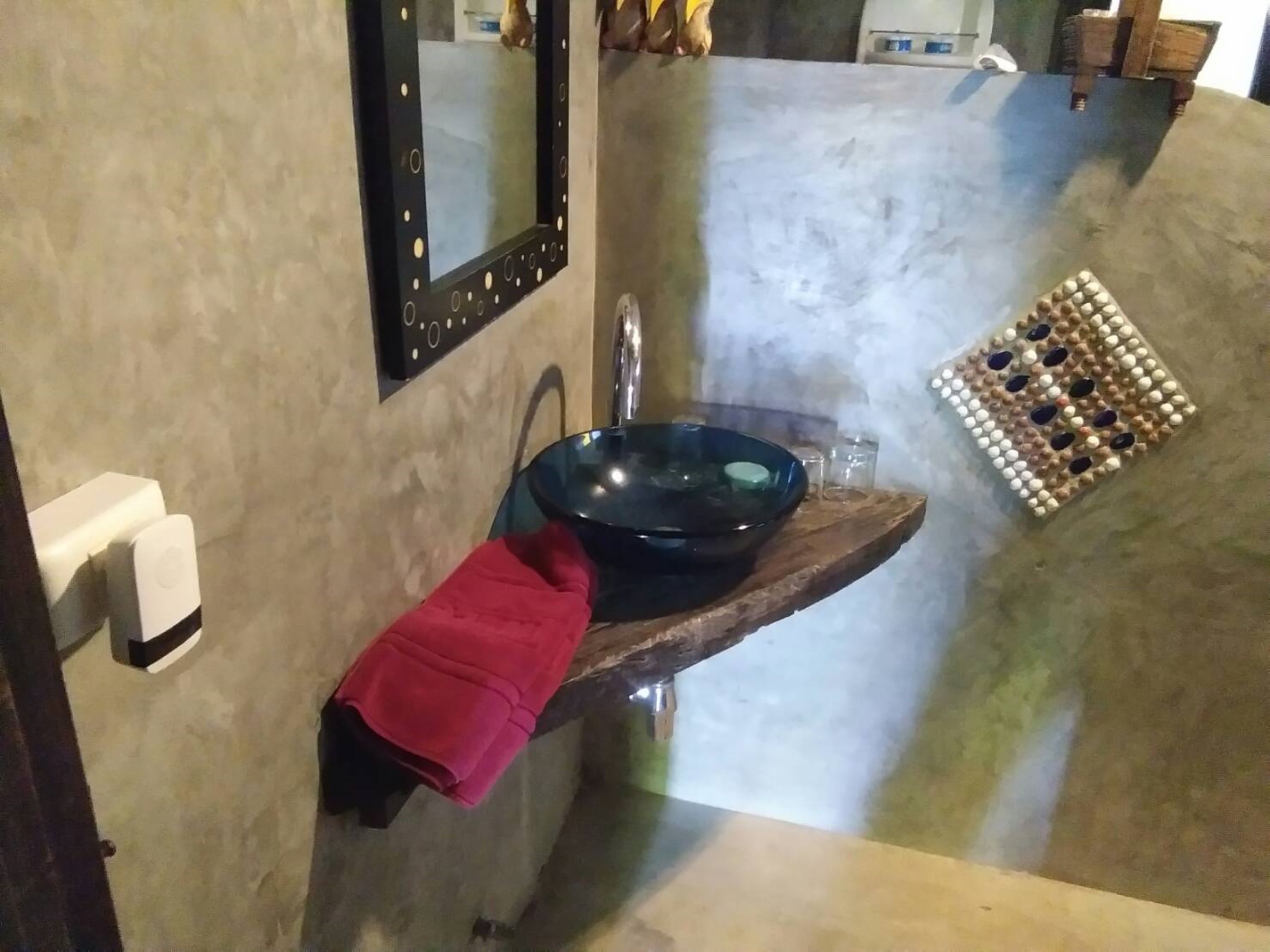 A bathroom with a round, dark blue glass basin on a wooden shelf, a red towel, a mirror with a black frame, and a small decorative item on the wall.