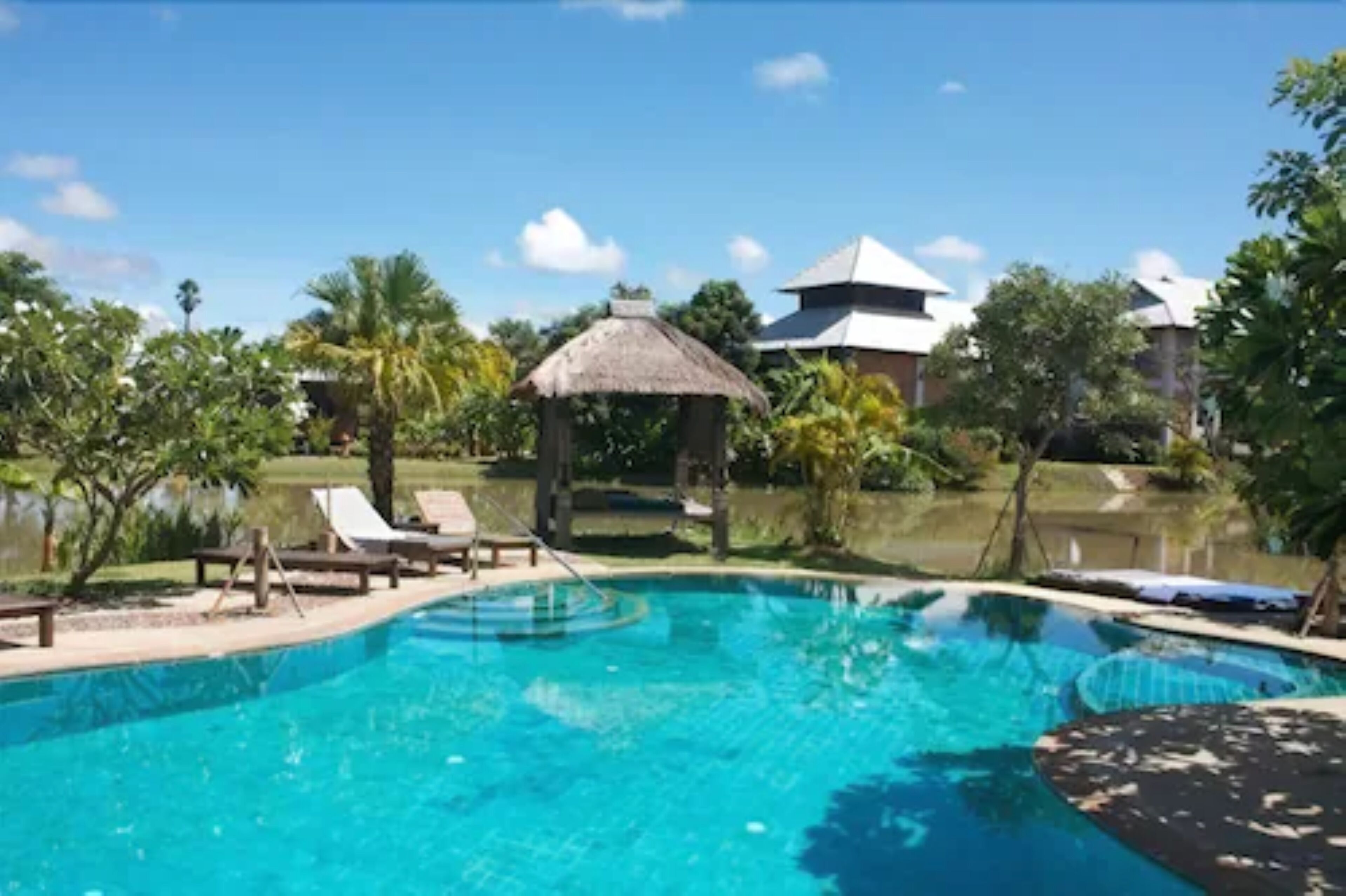 A clear blue pool with lounge chairs and a thatched structure, surrounded by tropical vegetation and buildings with white roofs.