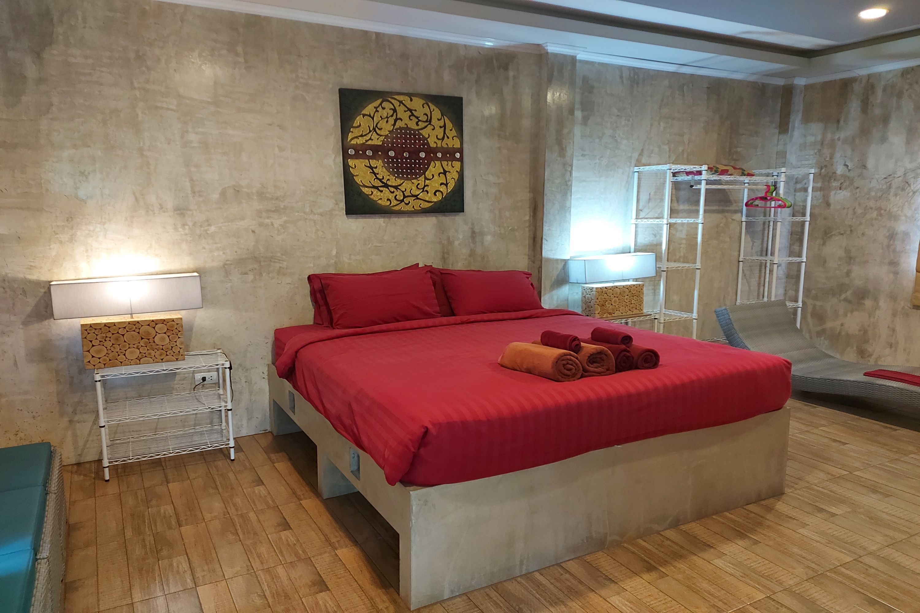 A bedroom with a large bed, red bedding, two bedside tables with lamps, a wall-mounted artwork, and a built-in shelf unit.