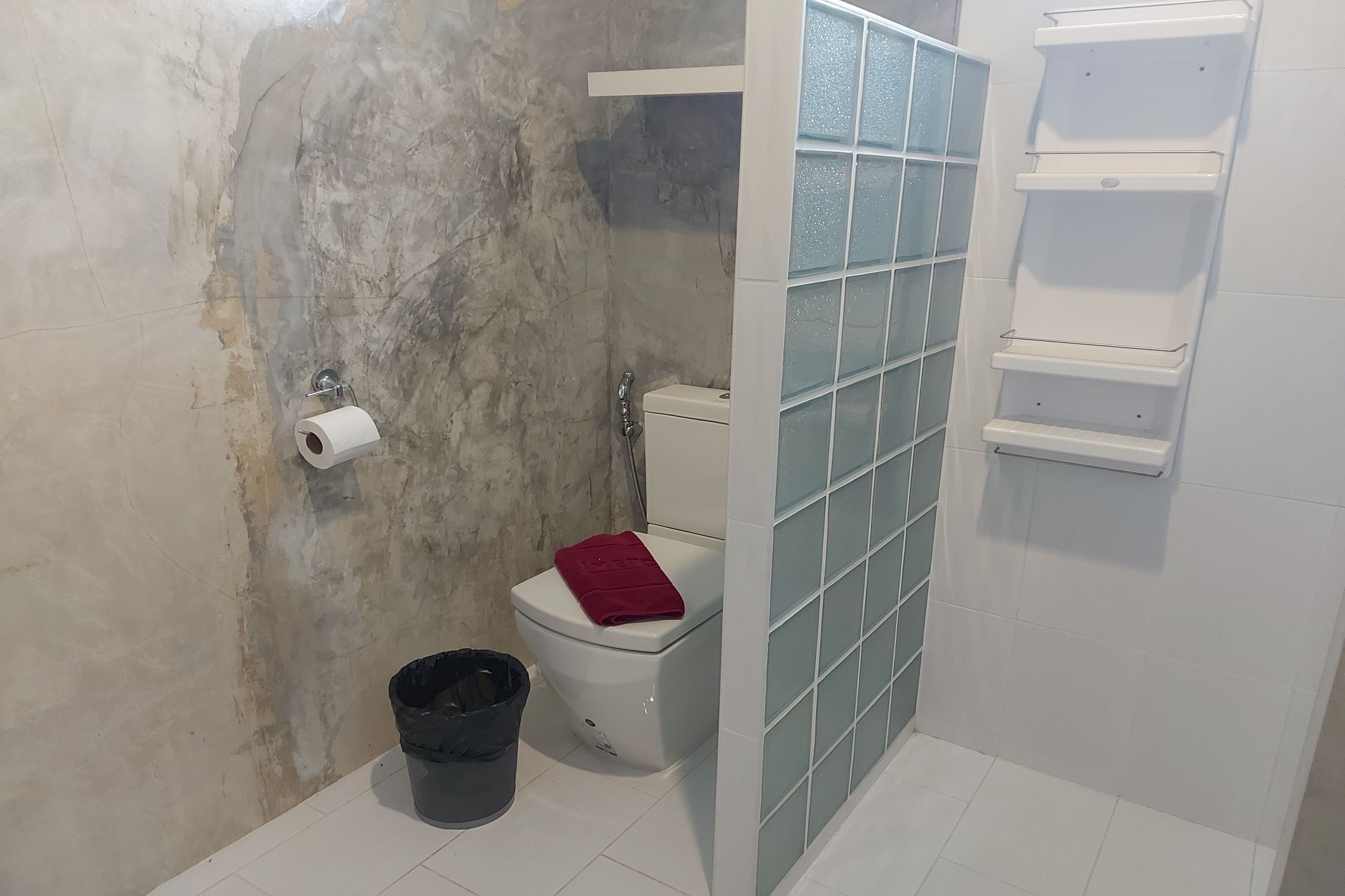 A bathroom with a toilet, a trash bin, and a shelf unit.