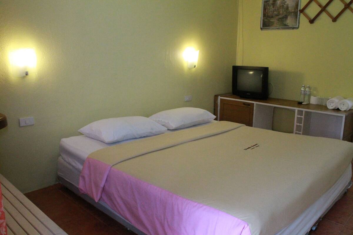 A single bed with a pink blanket, a television on a wooden stand, and a small table with toiletries.