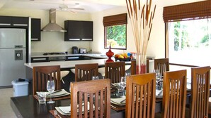 A modern kitchen with a central island, dining area with wooden chairs, and large windows with blinds. - Baan Chaba Samui (Koh Samui)