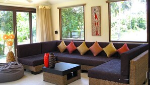 A modern living room with a dark-colored sofa, multiple cushions, a coffee table, and large windows offering a view of greenery outside. - Baan Chaba Samui (Koh Samui)