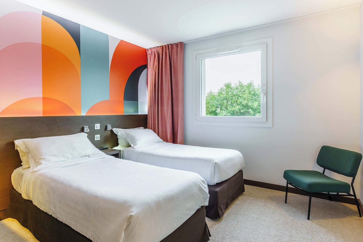 Ein Hotelzimmer mit zwei Betten, einem grünen Sessel und einem Fenster mit Blick auf Grünflächen.