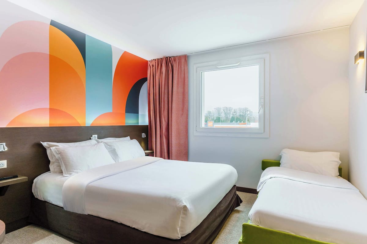 Ein Hotelzimmer mit einem großen Bett, einem kleineren Bett, einem Fenster und einem farbenfrohen Wandbild.