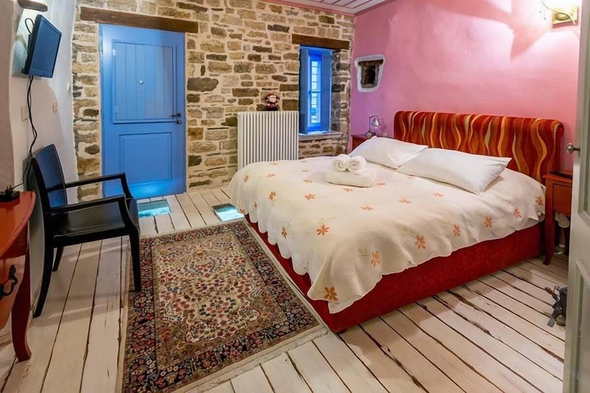 A bedroom with a stone wall, a blue door, a bed with a red headboard, and a patterned rug.