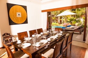 Un espace repas avec une longue table dressée pour un repas, entourée de chaises en bois. Une grande pièce décorative est accrochée au mur, et on aperçoit une piscine à travers une porte ouverte.