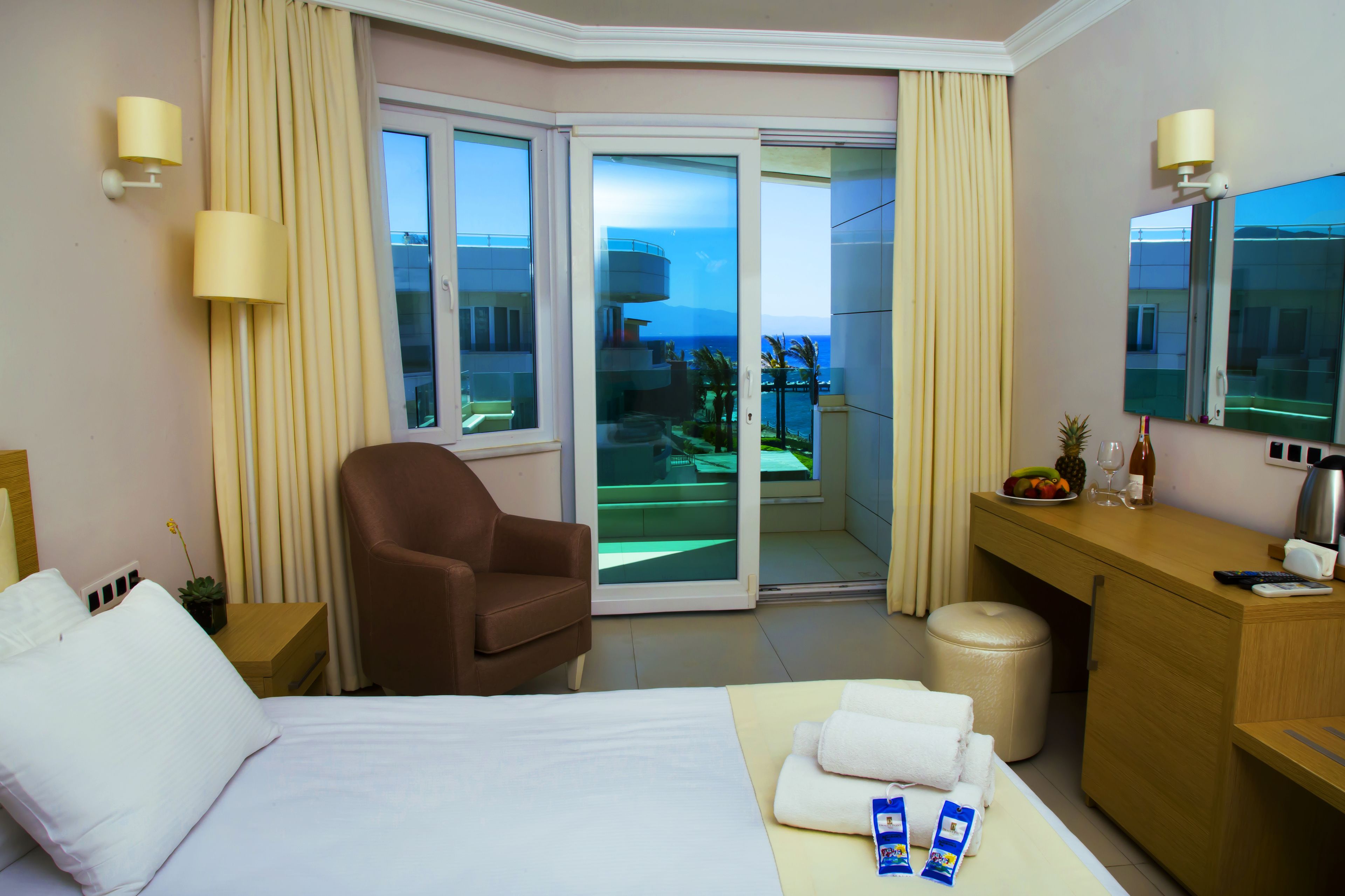 A hotel room with a bed, a brown armchair, a desk with a mirror, a small trash bin, and a view of the ocean through a sliding glass door.