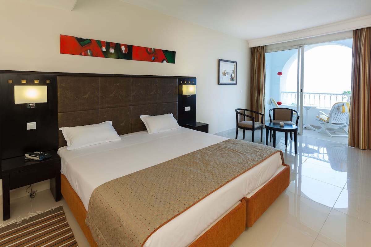 A hotel room with a large bed, two bedside tables, a nightstand, a chair, a small table, and a balcony with a chair and table.