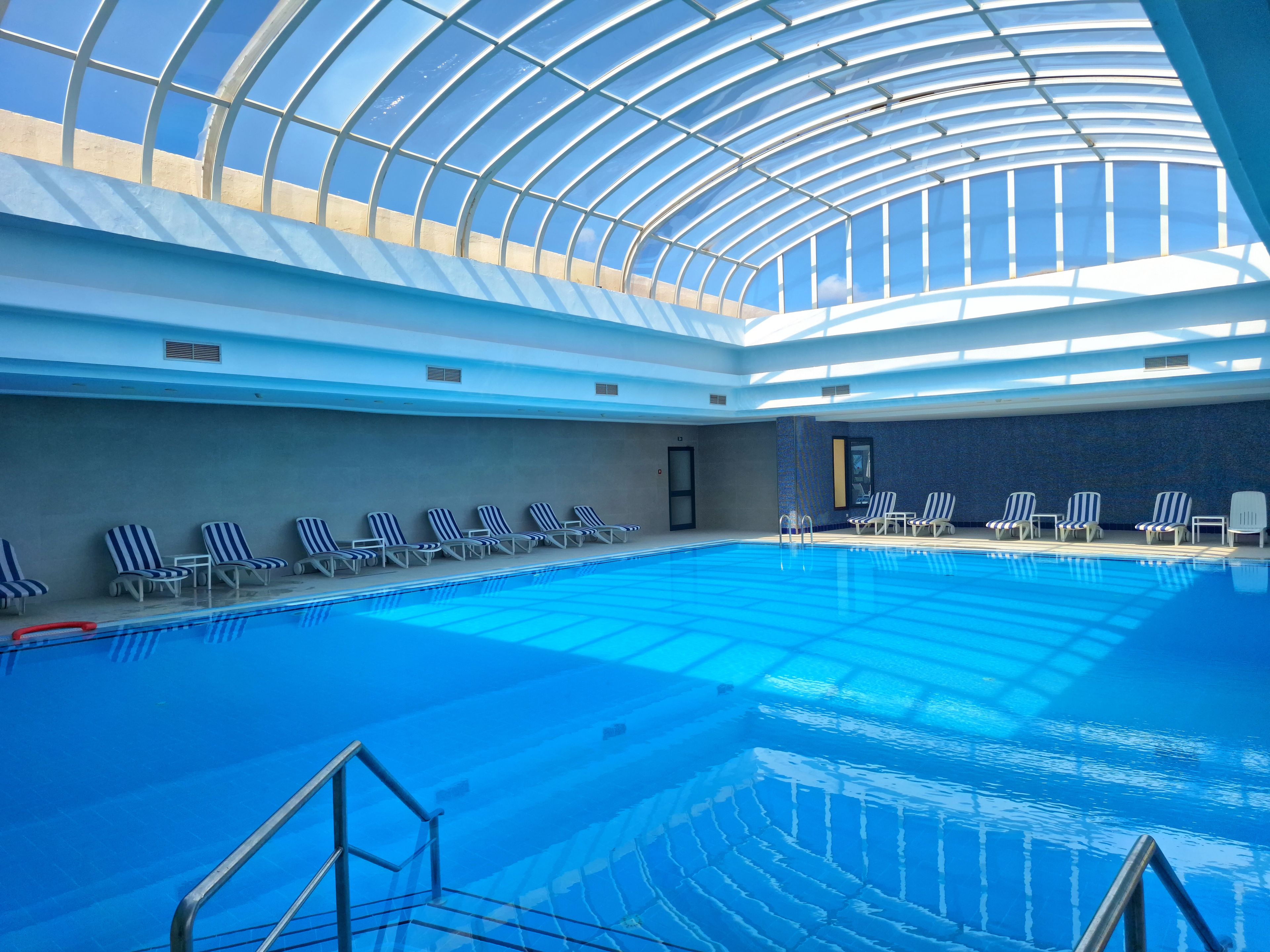 An indoor swimming pool with a glass ceiling, lounge chairs, and a tiled wall.